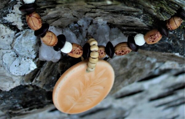 A close-up of the Bonnie necklace featuring a beaded design with brown, white, and tan beads and a round tan pendant engraved with a leaf, set against a textured stone background.